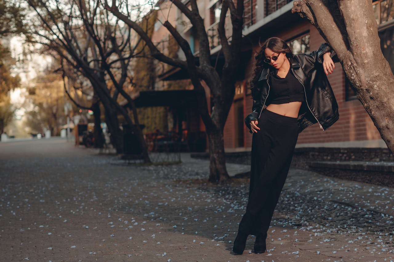 about-us-01 Fashionable woman in a black leather jacket posing stylishly on a tree-lined urban street.