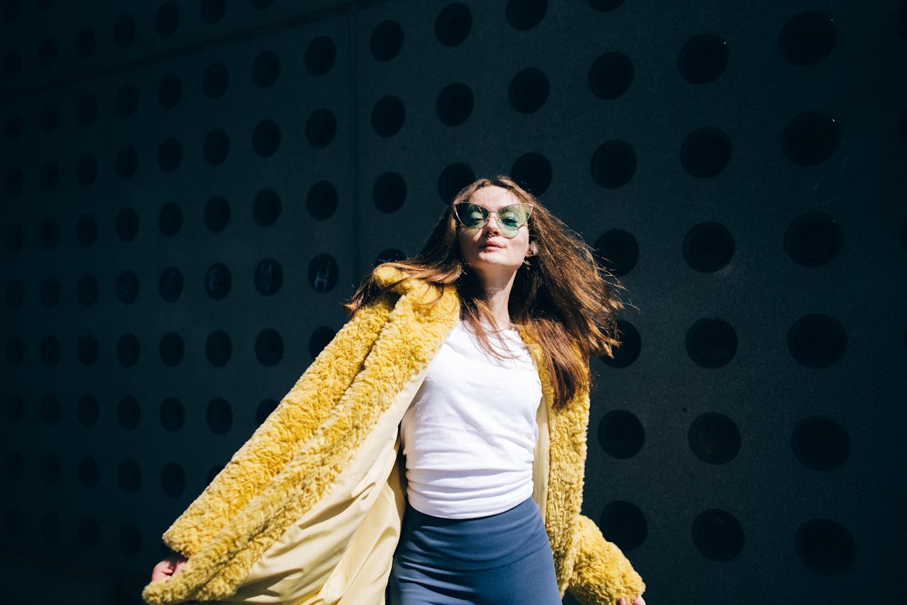 about-us-02 Confident woman wearing sunglasses and a yellow fur coat poses against a dotted wall in daylight.